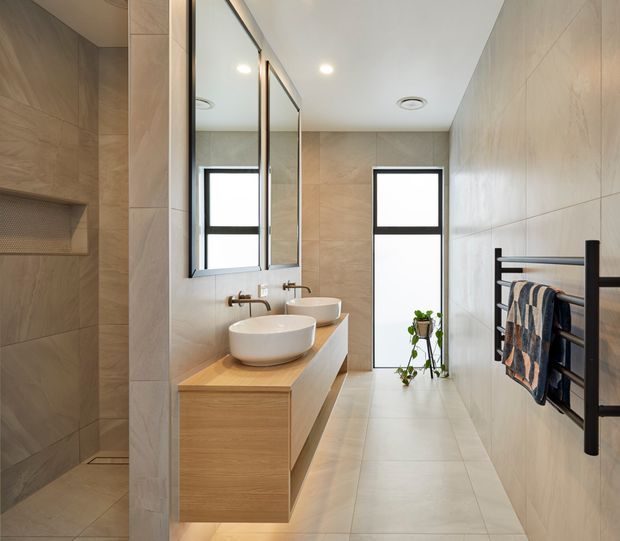 A contemporary bathroom with a long, narrow layout, large beige stone-look wall and floor tiles, a floating light-wood vanity with two vessel basins, wall-mounted brushed metal tapware, frameless mirrors, tall black-framed windows, and a black heated towel rail. A walk-in shower with a recessed niche is visible at left.