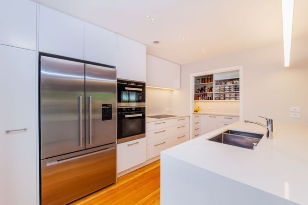 Bright contemporary white kitchen with extensive full-height cabinetry, a stainless-steel French-door refrigerator, integrated black appliances, a white island with double stainless-steel sinks, and warm timber flooring. A walk-in pantry with open shelving is visible at the rear.
