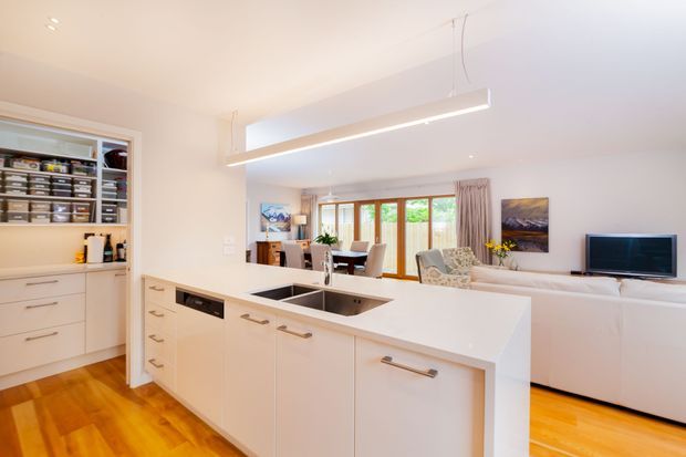 Bright open-plan kitchen and living area with a large white island, integrated sink and dishwasher, white built-in cabinetry, timber flooring, and a linear pendant light. A dining area and living room with sofa and television are visible beyond the kitchen, with timber-framed glazing bringing in natural light.