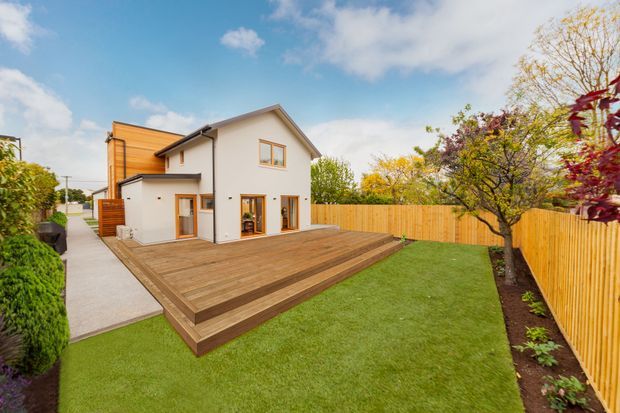 A bright rear exterior view of a two-storey suburban home with white rendered walls, warm timber cladding, a pitched gable roof, and large glazed doors opening onto a broad timber deck. The deck has stepped levels leading to a neatly landscaped lawn enclosed by a timber paling fence, with established garden planting around the perimeter.