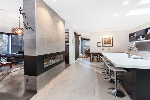 Open-plan contemporary kitchen and dining area adjoining a living room, centred around a large island with curved white benchtops and bar seating. A substantial tiled feature wall incorporates a linear fireplace, while recessed ceiling lighting, dark cabinetry, patterned splashback tile and full-height glazing create a refined modern interior.