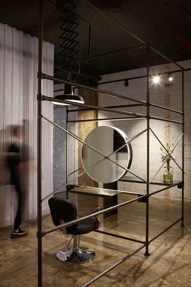 A moody industrial salon or styling space inside a converted warehouse-like interior. A black salon chair and circular mirror sit within a tubular metal scaffold framework, with exposed services, dark ceiling, white painted brick walls, polished concrete flooring, pendant and track lighting, and a translucent curtain partition. A small floral arrangement is visible in the background.