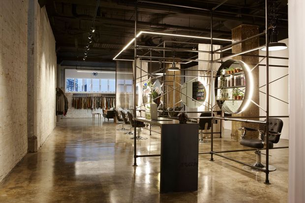 An industrial-style commercial interior, likely a fashion retail and styling space, with polished concrete flooring, exposed concrete and services, black metal shelving, illuminated circular display elements, clothing racks, salon-style chairs, and a central dark counter.