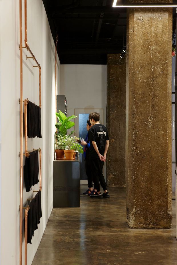 A narrow industrial-style interior corridor with polished concrete flooring, white walls, exposed copper pipework and black hanging textile elements on the left. Large rough concrete columns define the right side beneath a dark exposed ceiling. Potted plants and two people appear near a doorway at the end of the passage, with track and linear lighting overhead.