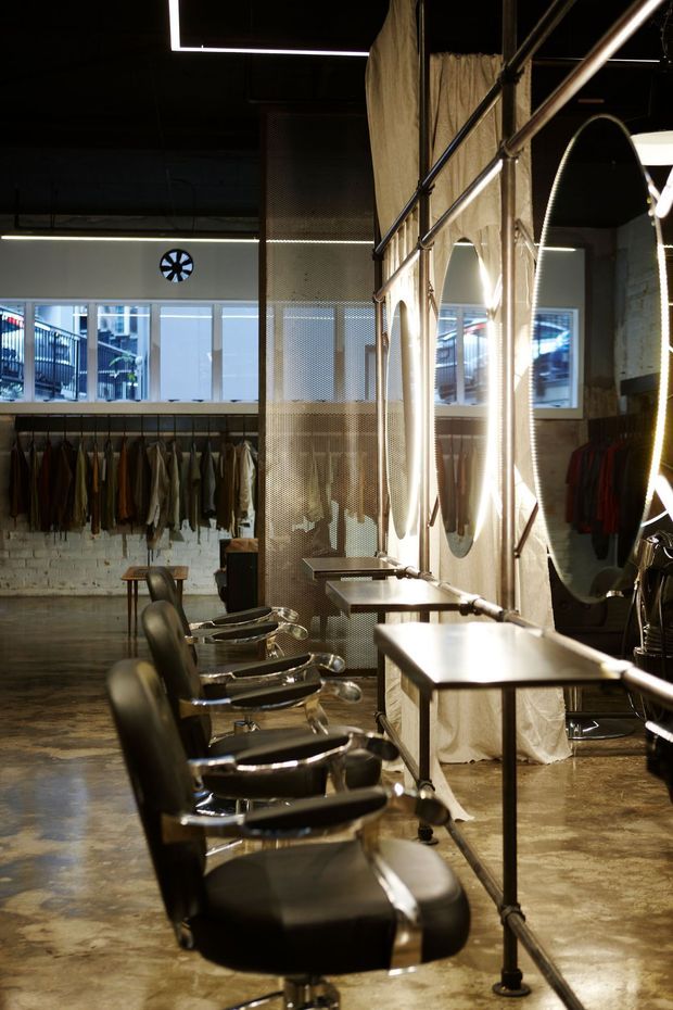 Industrial-style hair salon interior with a row of black salon chairs and styling stations, large mirrors framed by exposed metal piping, clothing or textile displays along the rear wall, polished concrete flooring, exposed white brick, mesh partitions, and dramatic linear lighting. The space has a dark ceiling and a raw, converted-commercial atmosphere.