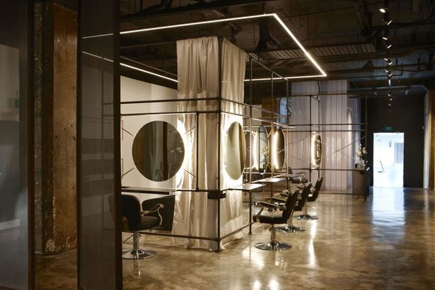 An expansive industrial-style interior, likely a hair salon or grooming studio, with polished concrete flooring, exposed services and structure, black metal framing, translucent mesh partitions, and curtained treatment or styling bays. Circular mirrors, salon chairs, and linear LED lighting define the open-plan space.