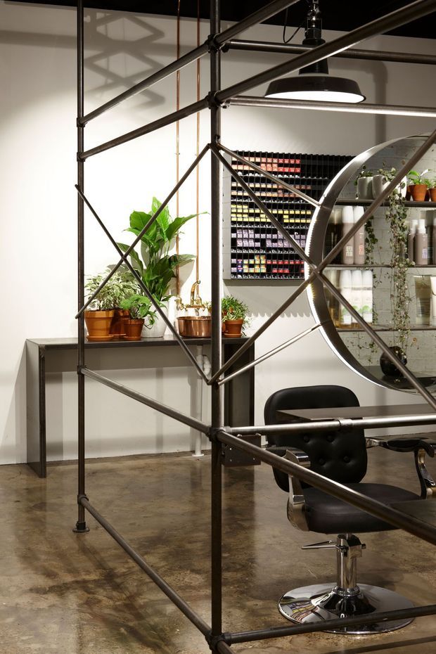 An industrial-style salon or retail workspace with exposed dark metal pipe scaffolding forming a prominent structural display frame. The space has polished concrete flooring, white walls, black track or pendant lighting, metal shelving with numerous small product containers, indoor plants, and a black leather barber or salon chair.