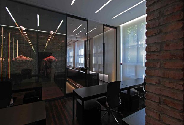 A dark, contemporary office interior with black workstations and chairs arranged beside full-height glass partitions. Exposed red brick forms a prominent wall, while linear recessed and suspended lighting creates strong reflections in the glazing. Window blinds provide filtered daylight, and the space has an industrial, moody character.