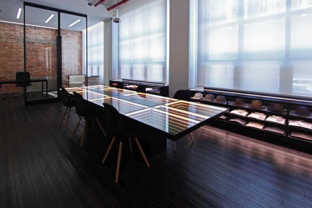 A dark, open-plan industrial-style dining and meeting space with a long glass-topped table surrounded by black chairs. Exposed red brick walls, black-framed glass partitions, large translucent windows, dark timber flooring, exposed ceiling services, and illuminated built-in display shelving define the room.