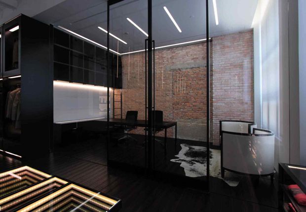 Dark, sophisticated dressing room or walk-in wardrobe with extensive black built-in joinery, illuminated shelving, glass partitions, a desk, and a lounge chair. Exposed red brick forms the feature wall, while linear LED lighting and dark flooring create an industrial-luxe atmosphere.
