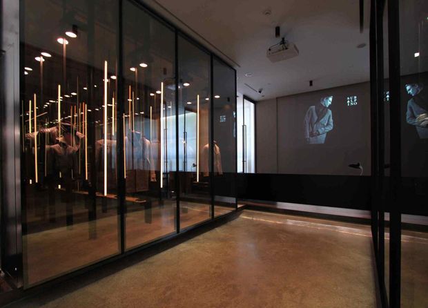 A dark, gallery-like commercial interior with black-framed glass display enclosures containing garments or merchandise, warm vertical pendant lighting, polished concrete flooring, and a projection on the far wall. The space has a minimalist, exhibition-oriented character with strong reflections in the glazing.