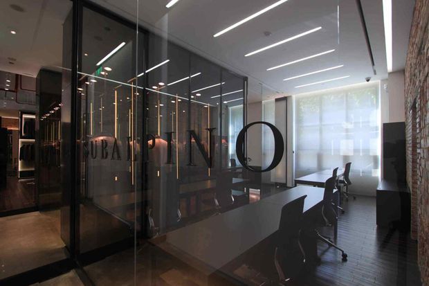 A contemporary commercial office interior with a glass-enclosed meeting room and large dark conference table. Linear recessed or suspended ceiling lights create strong horizontal lines, while a textured exposed brick wall contrasts with dark timber-look flooring, black-framed glazing, and dark office furniture. A reception or lounge area is visible beyond the glass partition.
