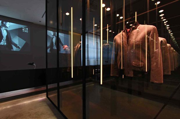 A dark, high-end retail or exhibition interior featuring a series of tailored jackets displayed behind black-framed glass partitions. Vertical warm light strips and repeated ceiling spotlights create reflections through the glazing, while a large monochrome fashion projection is visible on the rear wall. The space has a polished, dramatic, gallery-like presentation.