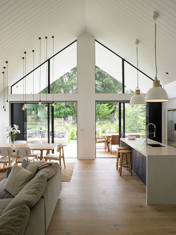 Bright open-plan kitchen, dining and living space with a dramatic pitched, white-lined ceiling and large triangular clerestory windows. Black-framed floor-to-ceiling glazing opens to a leafy garden and outdoor dining area. A long white stone island with dark cabinetry and pendant lights occupies the kitchen, while a timber dining table and pale upholstered sofa define the adjoining zones.