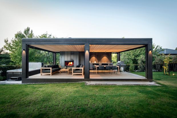 A contemporary open-sided backyard pavilion with a dark framed structure, timber-lined ceiling, concrete floor, and large glazed or open openings. The covered space is divided into a lounge area with seating and a fireplace, and an outdoor dining and cooking area with a table, chairs, and barbecue. It opens onto a landscaped lawn and garden in the evening.