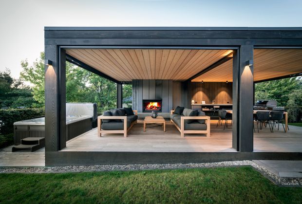A modern, open-sided outdoor entertaining pavilion with a dark timber-clad structural frame, warm timber ceiling, and concrete-look floor. The space includes a central fireplace, lounge seating, dining area, and an outdoor kitchen or barbecue zone, set within a landscaped garden.