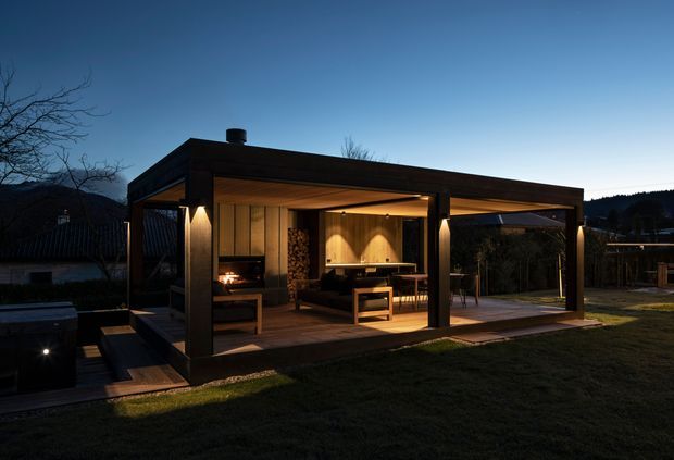 A freestanding contemporary backyard pavilion or outdoor entertaining structure photographed at dusk. The open-sided timber-clad shelter has a flat roof, substantial dark structural posts, warm recessed lighting, built-in seating and a dining table. A compact outdoor kitchen or barbecue area with stacked firewood is visible at the rear, while paving, lawn and surrounding fencing form the landscape setting.