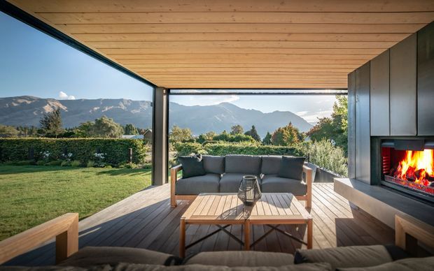 A covered outdoor lounge terrace with a timber-lined ceiling, timber decking, built-in seating and a central coffee table. A contemporary fireplace is integrated into the dark side wall, while the open sides frame landscaped grounds and mountain views.