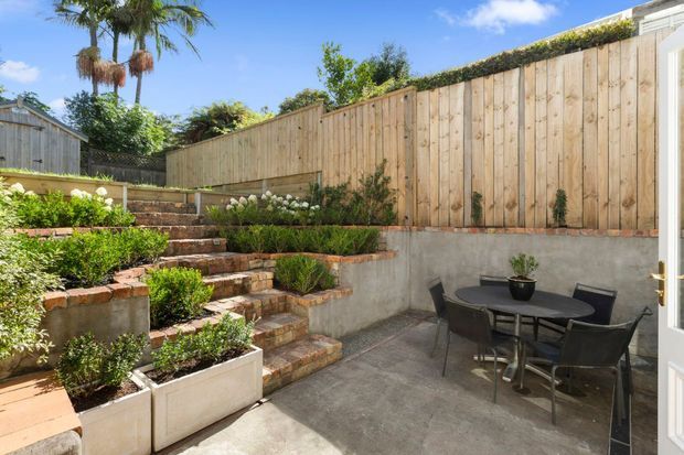 A sunny landscaped courtyard or patio with a paved outdoor dining area, black table and chairs, raised garden beds, terraced brick steps, rendered retaining walls, and a tall timber boundary fence. Tropical planting and a bright blue sky create a warm, private outdoor setting.