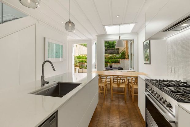 Bright, narrow contemporary kitchen with white full-height cabinetry, a long island with integrated stainless-steel sink, engineered stone benchtop, large stainless-steel range cooker, and a small pale mosaic-tile splashback. The kitchen opens into a timber-floored dining area with a timber dining table and chairs, abundant glazing, and a skylight. A planted courtyard is visible beyond the rear opening.
