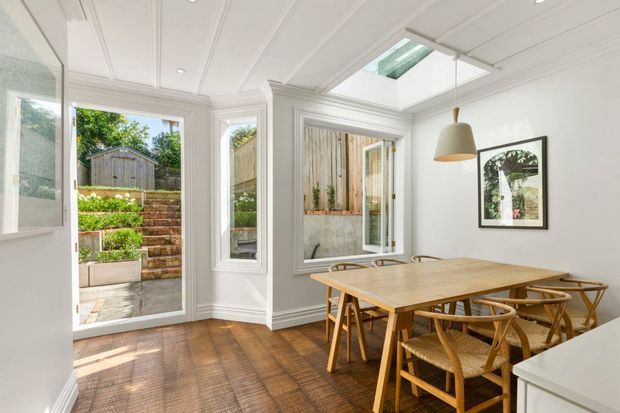 Bright dining room with a timber dining table and woven wooden chairs, white paneled walls and ceiling, timber flooring, framed artwork, and a large glazed door and windows opening to a landscaped courtyard. A skylight and pendant light provide additional illumination.