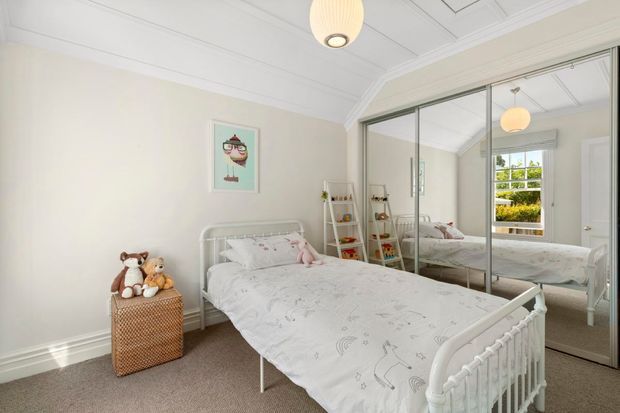 Bright, softly styled bedroom with a white metal bed, pale patterned bedding, mirrored sliding wardrobe doors, plush carpet, high decorative ceiling with panel moulding, and a small shelving unit with children's items. A window brings in natural light and greenery is visible outside.