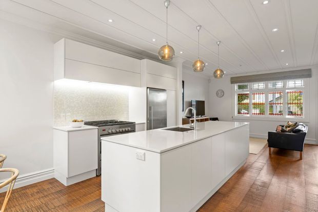 Bright, spacious open-plan kitchen with a large white island, integrated cabinetry, stainless-steel appliances, mosaic tile splashback, timber flooring, pendant lights, and a small adjoining living area with a window seat or lounge chair. The room features traditional detailing such as panelled ceilings, decorative trims, and a multi-pane window, combined with contemporary kitchen joinery.