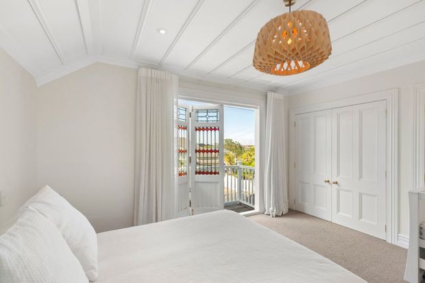 Bright, refined bedroom with a white upholstered bed, soft neutral carpet, full-height curtains, paneled white built-in wardrobe doors, and a decorative woven pendant light. French doors open to a small balcony with a scenic outlook, while the ceiling features decorative recessed panel detailing and traditional crown moulding.