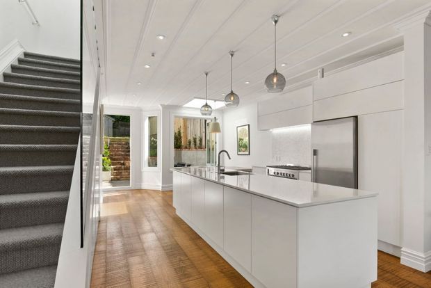 Bright contemporary open-plan kitchen viewed from beside a carpeted staircase. The space features a large white island with integrated sink, white handleless cabinetry, stainless-steel refrigerator and range, a pale tiled splashback, timber flooring, pendant lights, and white paneled ceiling detailing. Glazed doors and windows provide views toward a small outdoor courtyard.