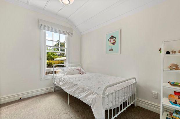 Bright, compact child’s bedroom with a white metal bed, pale bedding, soft neutral walls, carpeted floor, built-in open shelving filled with toys, framed playful artwork, and a large window with a roller blind. The room has a simple traditional architectural shell with a white paneled sloped ceiling and detailed skirting boards.
