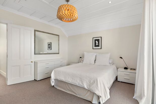 Bright, calm bedroom with a white upholstered bed dressed in white linen, matching white bedside tables and dresser, pale neutral walls, carpeted flooring, white panelled door, decorative wall moulding, and a large framed mirror. A woven timber pendant light adds a warm natural accent, while full-height white curtains soften the room.
