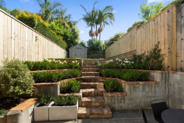 A sunny landscaped courtyard garden with a central run of rustic brick steps leading upward between terraced raised planting beds. Tall timber privacy fences line both sides, with clipped shrubs, flowering plants, and palms visible beyond. A small timber garden shed sits at the top of the steps, and part of an outdoor table and chair appears in the lower right.
