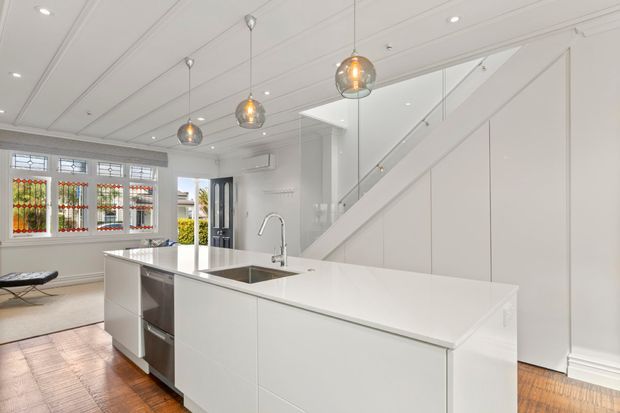 Bright open-plan kitchen with a large white island, integrated sink and curved chrome tap, pendant globe lights, white built-in cabinetry, and a glass-panelled staircase. Decorative leadlight-style windows and warm timber flooring add character to the otherwise clean, contemporary interior.