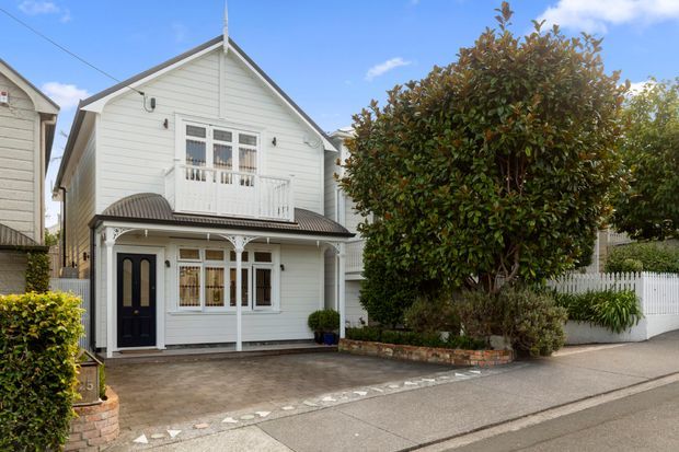 Street-facing view of a compact two-storey white weatherboard home with a steep gabled roof, decorative balcony and verandah, dark front door, paved driveway, and landscaped front garden enclosed by a white picket fence.