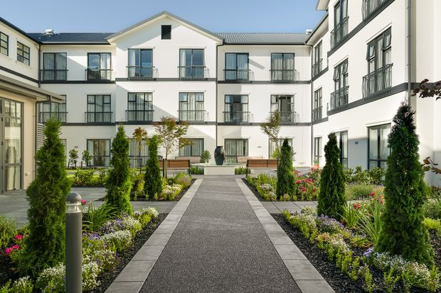 A landscaped communal courtyard framed by a multi-storey white residential building with dark metal roofing, evenly spaced windows, and black metal Juliet-style balconies. A straight paved pedestrian path runs through formal garden beds with clipped evergreen shrubs, flowering plants, and contemporary bollard lighting.