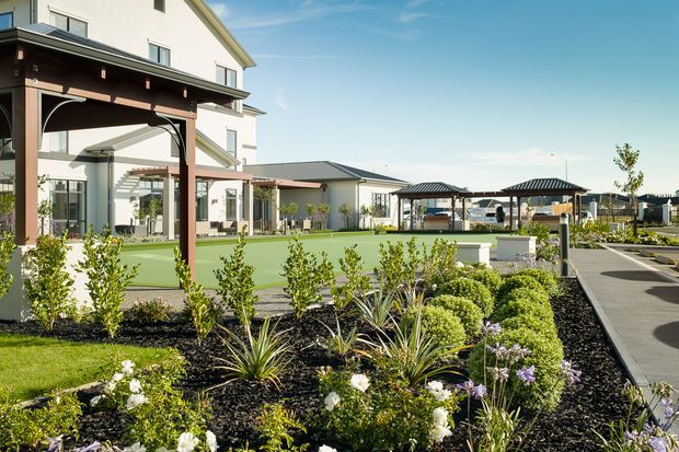 Landscaped outdoor communal area beside a large light-coloured multi-storey building, featuring a manicured garden with shrubs and flowering plants, a paved pedestrian path, covered timber-framed structures, and a green recreational court or lawn area. The architecture has a contemporary gabled form with dark metal roofing and white rendered walls.