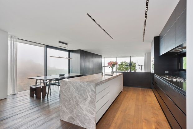 Contemporary open-plan kitchen with a large veined stone island, dark timber or matte cabinetry, integrated appliances, warm timber flooring, and an adjacent dining area. Full-height glazing and sheer curtains bring in abundant natural light, while linear recessed ceiling lighting reinforces the minimalist design.
