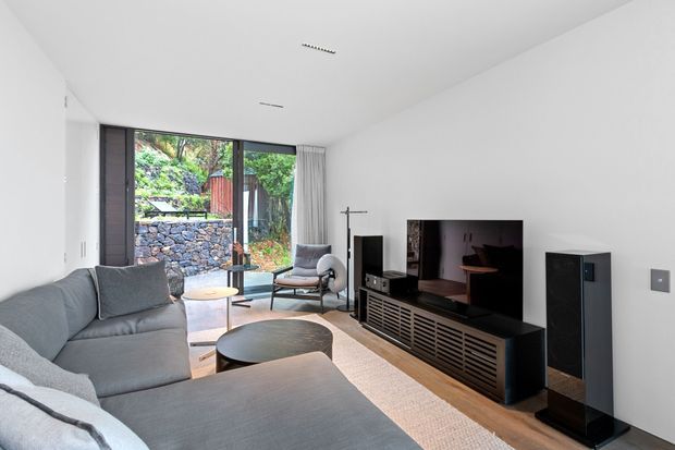 A bright, minimalist living room with a large grey sectional sofa, round coffee table, freestanding lounge chair, and a wall-mounted television on a dark slatted media console. Full-height glazing and a glass door open to a landscaped stone garden, while white walls, light timber flooring, and concealed ceiling lighting create a clean contemporary interior.