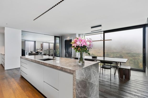 Contemporary open-plan kitchen and dining area featuring a large waterfall island with grey-veined stone, sleek white handleless cabinetry, timber flooring, black-framed floor-to-ceiling glazing, and a minimalist dining table. The space is bright and refined, with expansive views through the windows.