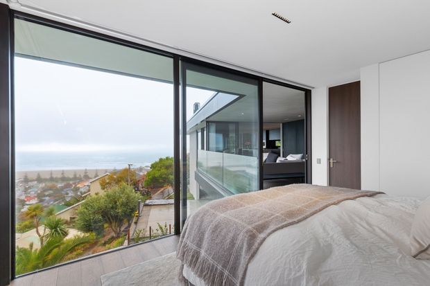 A contemporary bedroom with a neatly made bed positioned beside expansive floor-to-ceiling black-framed glazing. The windows overlook a coastal residential landscape and ocean, while a glazed balcony or terrace and an adjoining modern interior are visible beyond. The room has white walls, a dark timber door, recessed ceiling lighting, and a restrained neutral palette.