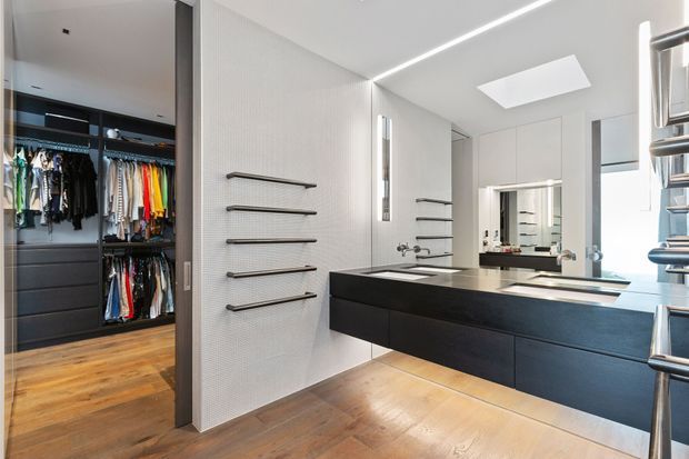 Contemporary bathroom with a floating dark vanity, integrated double basins, large mirror, wall-mounted towel rails and a skylight. An open walk-in wardrobe with hanging clothes and drawer storage is visible to the left, while warm timber flooring contrasts with the crisp white wall surfaces and black joinery.