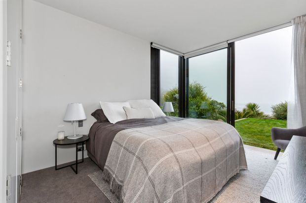 Bright contemporary bedroom with a neatly made bed, grey checked throw, dark bedside table, and table lamps. Full-height corner glazing opens views to greenery and an expansive outdoor landscape, while white walls and sheer curtains create an airy feel.