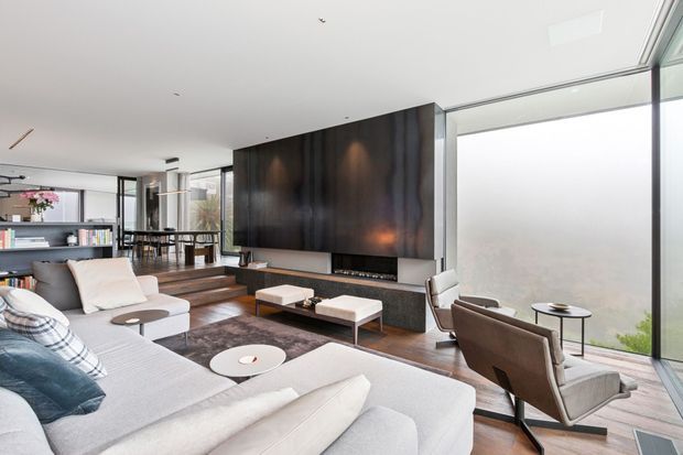 Contemporary open-plan living room with a large pale sectional sofa, sculptural round coffee tables, brown lounge chairs, and a broad dark feature wall incorporating a fireplace and television. Full-height glazing provides abundant natural light, while timber flooring, recessed lighting, and a partially visible dining area create a refined, spacious interior.