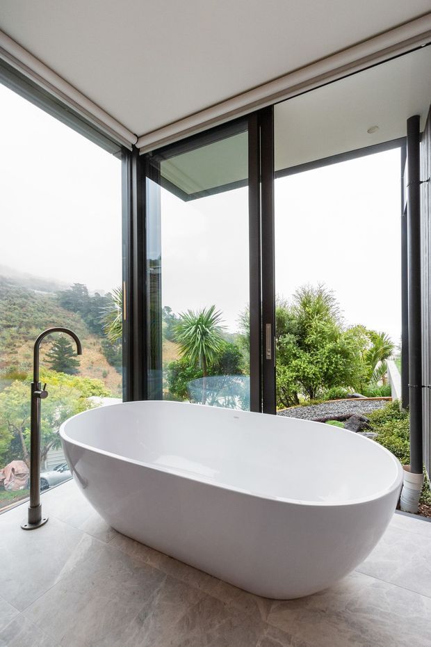 A bright, contemporary bathroom featuring a large freestanding white oval bathtub and floor-mounted brushed metal faucet. Full-height dark-framed glazing wraps around the corner, opening to lush landscaped greenery and hillside views. The floor is finished in pale stone-look tiles, with a clean, spa-like atmosphere.