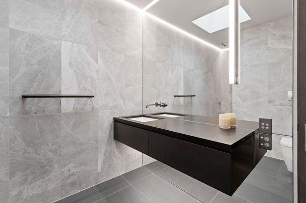 Contemporary bathroom featuring large-format grey marble-look wall tiles, dark grey floor tiles, a floating black vanity with an integrated light grey benchtop and twin basins, wall-mounted fixtures, a large mirror, recessed linear LED lighting, and a skylight. A toilet is visible at the right.