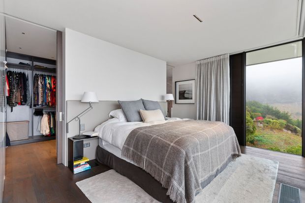 Contemporary bedroom with a neatly made upholstered bed, layered grey and white bedding, bedside tables, wall-mounted reading lights, framed artwork, full-height curtains, and a walk-in wardrobe visible at left. Dark timber flooring and a large glazed opening provide a refined connection to the landscaped exterior.