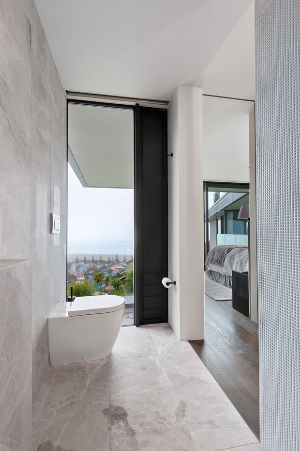 A contemporary bathroom with a wall-mounted white toilet, pale marble-look tiled walls and floor, and a dark-framed glazed door opening to an exterior view. A bedroom with timber flooring and a bed is visible directly through the open doorway, establishing the bathroom as an ensuite. A patterned translucent screen occupies the right side, while the bathroom overlooks a coastal urban landscape.
