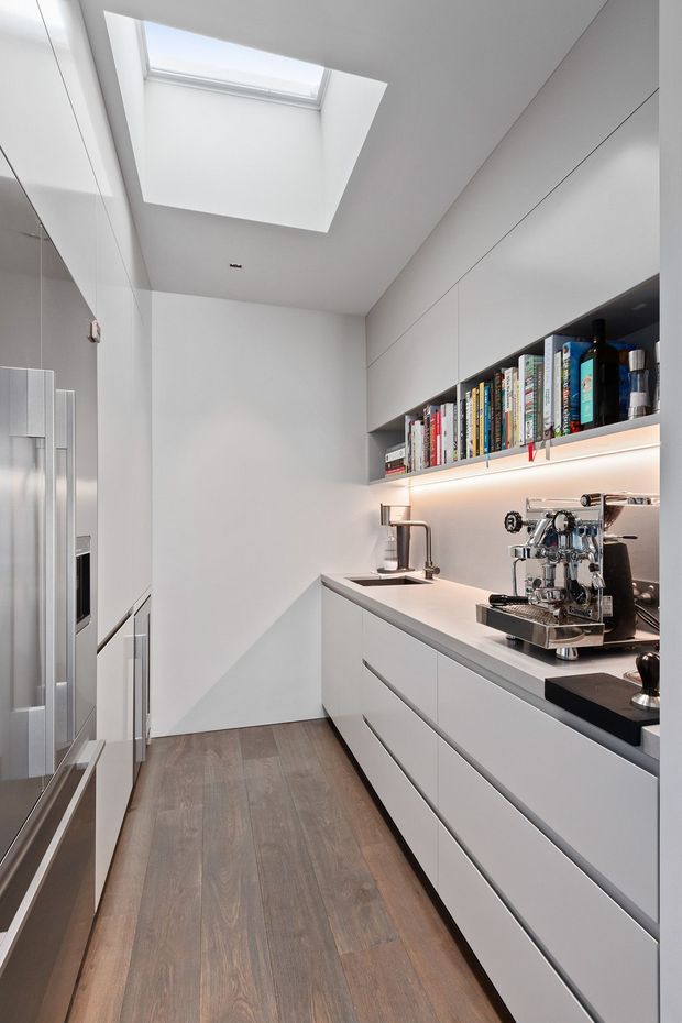 A narrow, highly minimalist kitchen or kitchenette with full-height white cabinetry, integrated appliances, a long white benchtop, undermount sink, open illuminated shelving with books, and a polished espresso machine. A large skylight brings natural light into the space, while the warm timber-look floor adds contrast to the predominantly white fit-out.