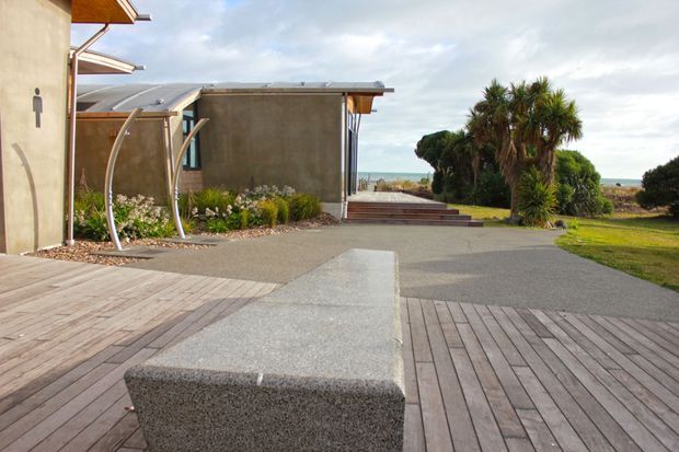 Exterior view of a contemporary coastal property, showing a broad timber deck, textured concrete-paved courtyard, built-in concrete bench, curved metal outdoor structure, low flat-roofed building volumes, and landscaped planting with the sea visible beyond.
