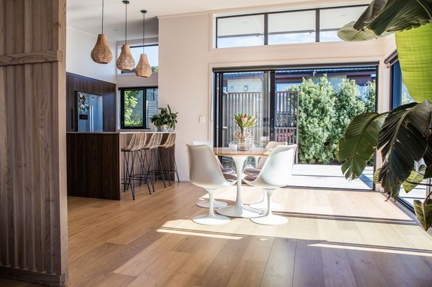 Bright open-plan dining and kitchen area with warm timber flooring, a white tulip-style dining table and chairs, dark timber kitchen cabinetry, woven pendant lights, and large glazed doors and windows opening to a planted outdoor area. A timber-clad vertical feature wall frames the left side, while large indoor plants add a tropical accent.